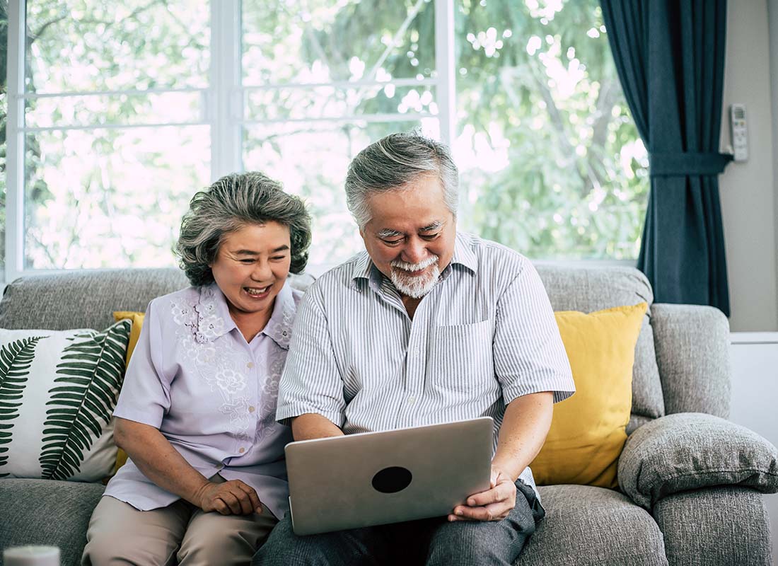 General-Medicare-Senior-Couple-Laughing-While-Sitting-on-the-Sofa-in-a-Bright-Living-Room-and-Using-Laptop-to-Review-Their-Medicare-Options